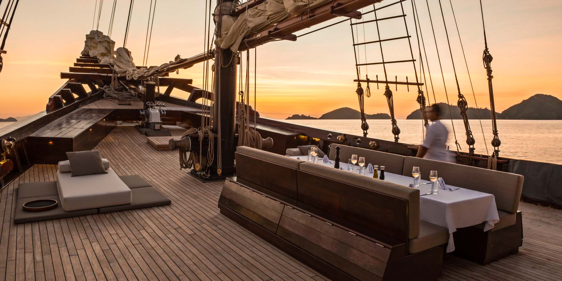 A crew member walking past a fine dinner setup on the expansive foredeck of Dunia Baru during sunset