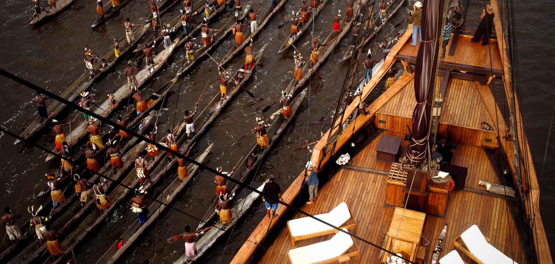 A fleet of Asmat rowing wooden canoes with warriors in traditional dresses greeting Silolona on arrival at Syuru village, Agats