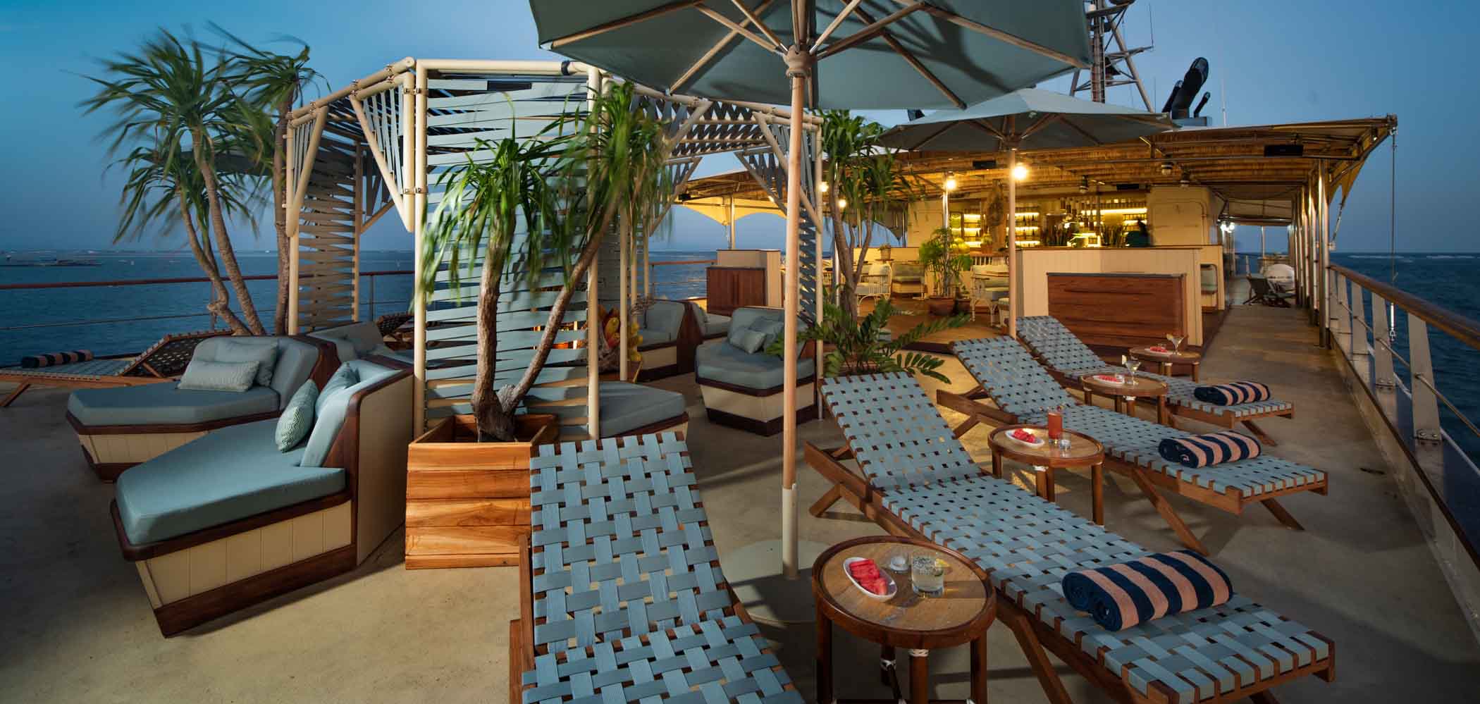 Outside relaxing lounge area with palm trees, sofas and beds on the top deck of the Kudanil Explorer superyacht at dusk